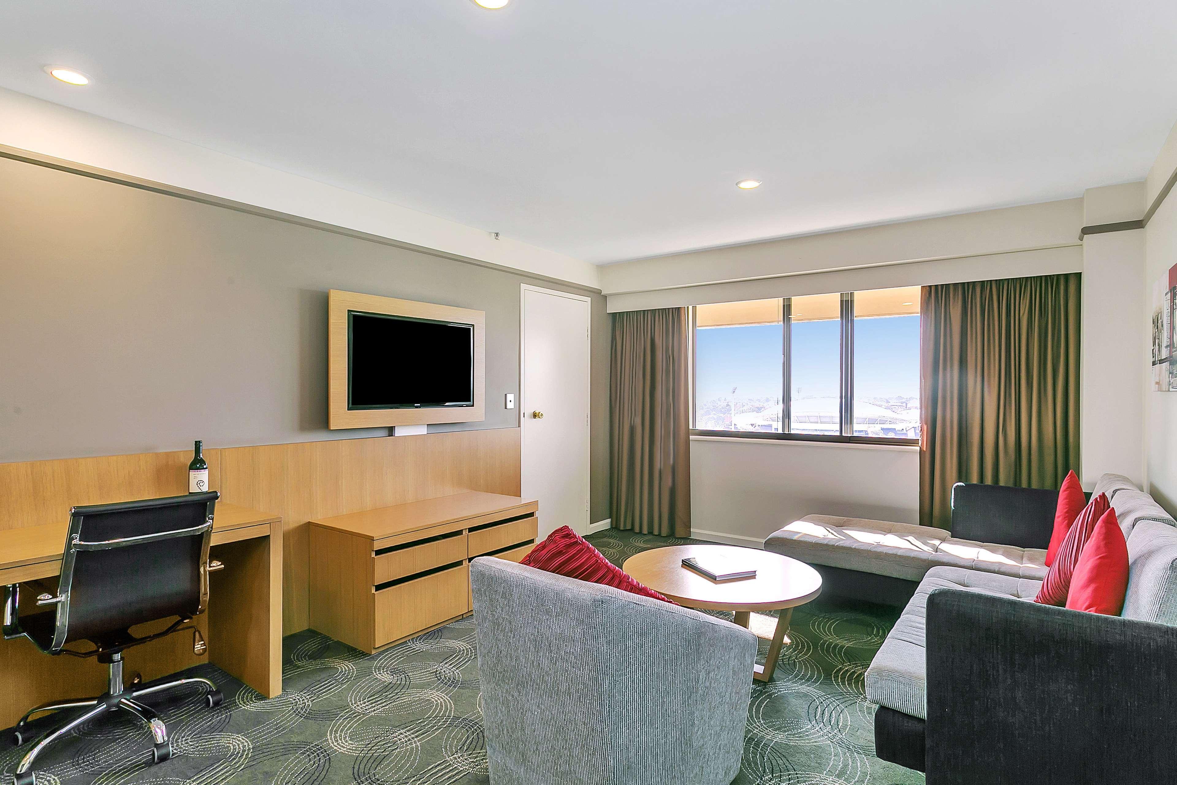 Hotel Stamford Plaza Adelaide Exteriér fotografie The photo shows a modern hotel room or suite that features a cozy seating area. There is a comfortable sofa with red accent pillows, a round coffee table in the center, and a sleek desk with an office chair. On the wall, there is a flat-screen TV mou