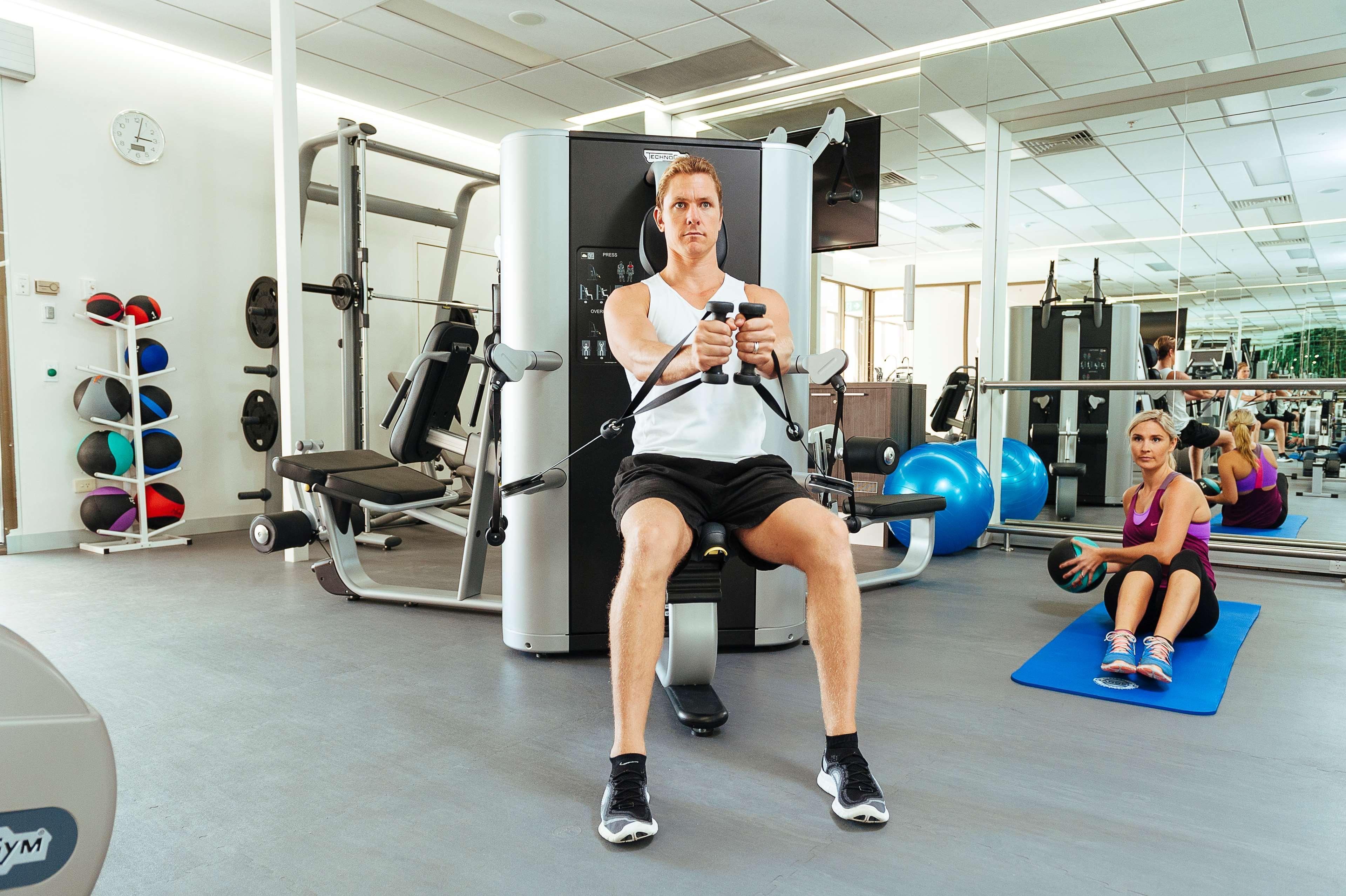 Hotel Stamford Plaza Adelaide Exteriér fotografie The photo shows a gym setting with two individuals engaged in workout activities. 

In the foreground, a man is seated on a machine, using the handles to perform a rowing exercise. He is wearing a sleeveless shirt and shorts, indicating an active wor