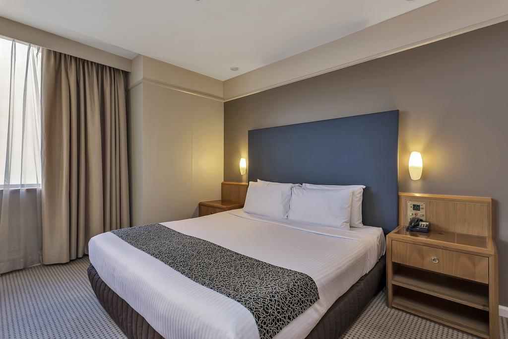 Hotel Stamford Plaza Adelaide Exteriér fotografie The photo shows a modern hotel room. It features a double bed with a white duvet and a decorative grey and black throw. The headboard is a solid dark color. There are two bedside tables on either side of the bed, each with a lamp. Natural light enter